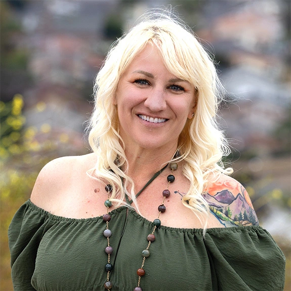 Photograph of Brooke York smiling brightly in natural light, wearing a cheerful outfit with long blonde hair, cityscape softly visible in the background.
