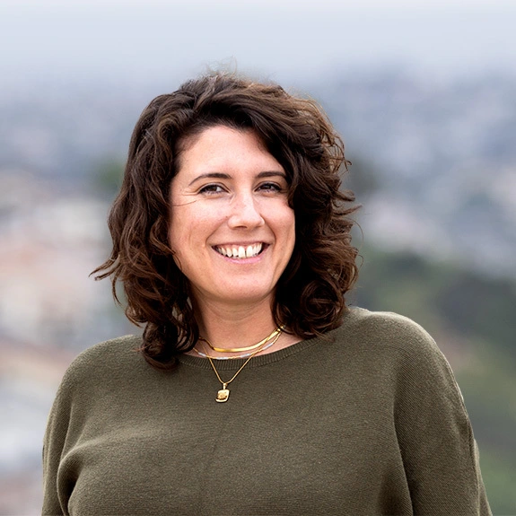 Photograph of Jessica Cyr smiling outdoors with curly hair and a casual outfit, set against a blurred city backdrop