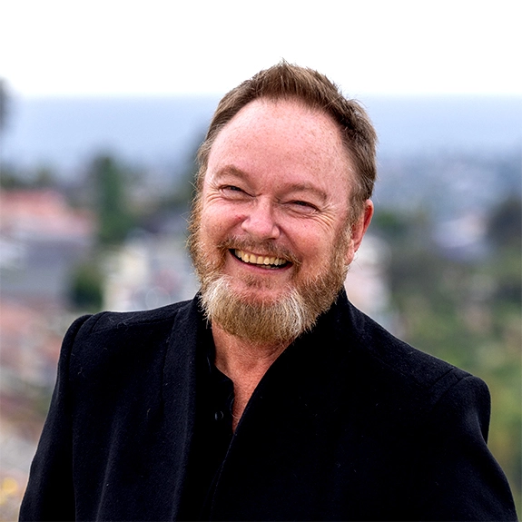 Photograph of Max Parker smiling outdoors in a black jacket, with a blurred hillside cityscape in the background.
