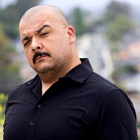Photograph of Ricky Perez outdoors in a dark jacket, looking confidently toward the camera with a blurred city view behind him.